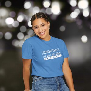 May include: A sky blue t-shirt featuring the text "ACCOUNTABILITY IS NOT OPTIONAL" in a white rectangular box. The shirt is paired with blue denim jeans. The background has blurred bokeh lights.
