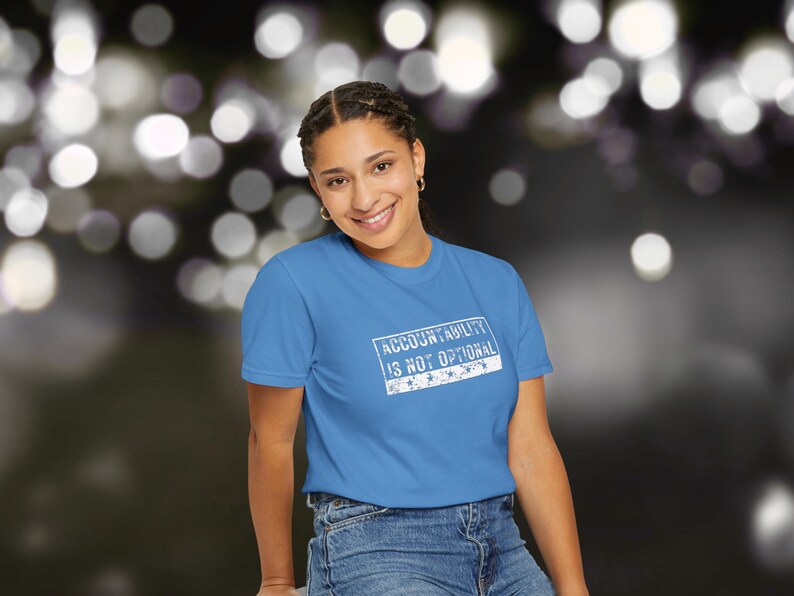 May include: A sky blue t-shirt featuring the text "ACCOUNTABILITY IS NOT OPTIONAL" in a white rectangular box. The shirt is paired with blue denim jeans. The background has blurred bokeh lights.
