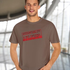 May include: A brown t-shirt featuring the text "ACCOUNTABILITY IS NOT OPTIONAL" in a red, distressed stamp design. The t-shirt is plain and has a crew neck.