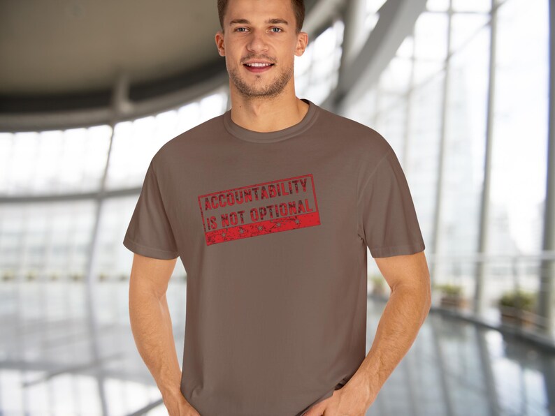 May include: A brown t-shirt featuring the text "ACCOUNTABILITY IS NOT OPTIONAL" in a red, distressed stamp design. The t-shirt is plain and has a crew neck.