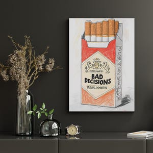 May include: A framed artwork depicts a pack of cigarettes with the words "BAD DECISIONS" in bold letters. The pack is red and white, with the cigarettes visible inside. The artwork is a humorous commentary on life choices.
