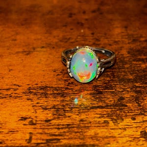 Puede incluir: Un anillo de plata con una piedra preciosa de ópalo ovalada. El ópalo muestra un vibrante juego de colores, incluyendo tonos verdes, azules y rojos. El anillo está sobre una superficie de madera, con un fondo borroso.
