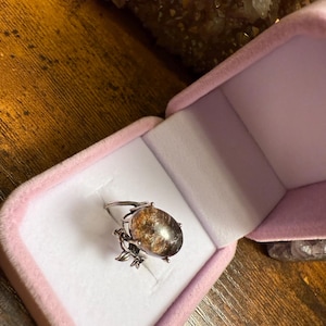 May include: A silver ring with a large, oval-shaped, brown and gold gemstone is displayed inside a pink velvet ring box. The ring has a decorative floral design on the sides of the gemstone.