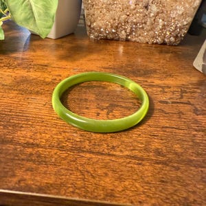 Może przedstawiać: Jasnozielona bransoletka typu bangle. Bransoletka to solidna, okrągła opaska o gładkim, błyszczącym wykończeniu. Bransoletka spoczywa na ciemnobrązowej drewnianej powierzchni.