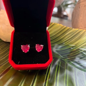 May include: A pair of heart-shaped ruby earrings displayed in an open red jewelry box with a black velvet interior. The earrings are set in silver or white gold. The background features a green and yellow leaf pattern.