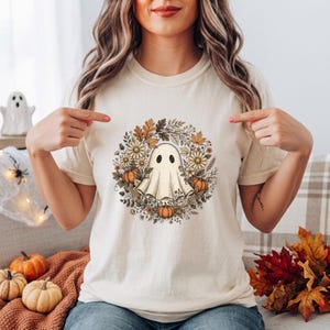 Peut inclure: Un t-shirt à manches courtes de couleur crème avec un motif central représentant un fantôme blanc entouré d'une couronne circulaire de feuilles d'automne, de citrouilles orange et de fleurs blanches imprimé sur le devant du vêtement.