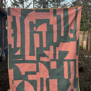 May include: A handmade quilt with a geometric design. The quilt features a pattern of dark teal shapes against a coral background. The edges are finished with a yellow binding. The quilt is held up outdoors.