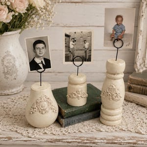 May include: Three cream-colored photo holders with black wire stems, each holding a vintage photograph. The holders have ornate, raised floral designs. A vase of pink roses and antique books are in the background, creating a vintage aesthetic.