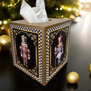 May include: A decorative tissue box with a gold frame and black panels featuring illustrations of nutcracker soldiers. The box has a checkered pattern on the edges and is filled with white tissues. Gold ornaments and a Christmas tree are in the background.
