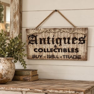 May include: A rectangular wooden sign with a distressed white finish and black lettering that reads Antiques & Collectibles Buy - Sell - Trade. The sign features a decorative carved molding at the top and hangs from a thick jute rope.