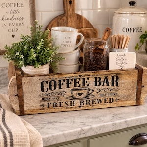 May include: A rectangular wooden box with metal accents featuring the black text COFFEE BAR, FRESH BREWED, OPEN 24/7, and SELF SERVE. The box contains a potted green plant, three stacked white mugs, a glass jar of coffee beans, and napkins.