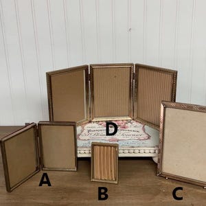 May include: A collection of antique bronze-colored picture frames in various sizes and styles. The frames include single, double, and triple folding designs, with decorative edges and cardboard backing. The frames are arranged on a wooden surface.