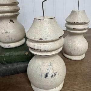 K&ouml;nnte beinhalten: Drei cremefarbene, rustikale Holz-Fotohalter mit Craquel&eacute;-Finish. Jeder Halter hat eine dekorative, gedrehte Form und eine Drahtklammer aus Metall zum Pr&auml;sentieren von Fotos oder Notizen. Die Halter stehen auf einer Holzoberfl&auml;che.