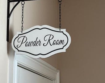 May include: A white, decorative sign with the words "Powder Room" in elegant script, framed by a black border. The sign hangs from a black metal bracket and chain, mounted above a white door.