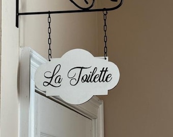 May include: A white sign with the words "La Toilette" in black script hangs from a black metal bracket and chain. The sign is cloud-shaped and mounted above a white door.