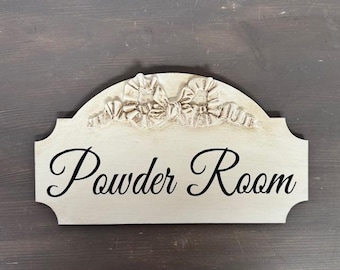 May include: Cream-colored wooden sign with the words "Powder Room" in elegant black script. The sign has a decorative, arched top with carved floral accents. The background is a dark gray.