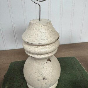 K&ouml;nnte beinhalten: Ein cremefarbener Foto- oder Notizhalter im Shabby-Chic-Stil mit Craquel&eacute;-Finish. Der Halter hat einen dekorativen, gedrechselten Holzsockel und eine silberne Drahtspirale. Der Halter steht auf einem gr&uuml;nen Buch.