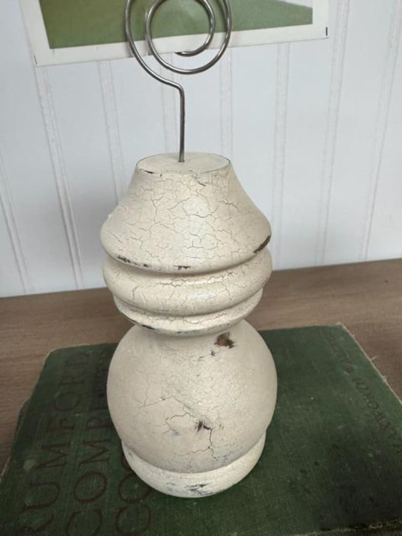 K&ouml;nnte beinhalten: Ein cremefarbener Foto- oder Notizhalter im Shabby-Chic-Stil mit Craquel&eacute;-Finish. Der Halter hat einen dekorativen, gedrechselten Holzsockel und eine silberne Drahtspirale. Der Halter steht auf einem gr&uuml;nen Buch.