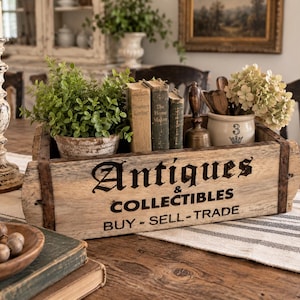 May include: A rectangular wooden crate with metal corner brackets featuring the black printed text Antiques & COLLECTIBLES BUY - SELL - TRADE. The crate contains a potted green plant, three vintage books, a metal bell, and a ceramic jar.