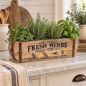 May include: A rectangular wooden planter box with dark metal accents, featuring the black printed text "HAND PICKED FRESH HERBS ORGANIC OPEN DAILY" and "OREGANO ROSEMARY BASIL THYME". The box is filled with green oregano, rosemary, thyme, and basil plants.