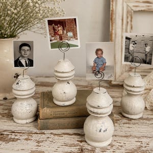 K&ouml;nnte beinhalten: Vier wei&szlig;e, shabby-chic Foto-Halter mit Drahtclips, die jeweils ein Vintage-Foto halten. Die Halter haben einen bauchigen Sockel und eine abgestufte Oberseite. Die Fotos zeigen Menschen und Szenen.