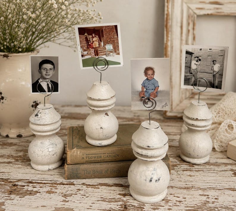 K&ouml;nnte beinhalten: Vier wei&szlig;e, shabby-chic Foto-Halter mit Drahtclips, die jeweils ein Vintage-Foto halten. Die Halter haben einen bauchigen Sockel und eine abgestufte Oberseite. Die Fotos zeigen Menschen und Szenen.