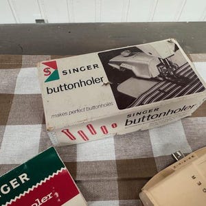 May include: Vintage Singer buttonholer box with the Singer logo and the words "buttonholer" and "makes perfect buttonholes". The box is white with red and green accents. Also visible are parts of other Singer sewing machine accessories.