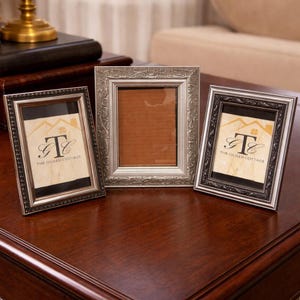 May include: Three decorative picture frames in silver and black, arranged on a dark wood surface. The frames have ornate detailing and include the text "THE GILDED COTTAGE". The frames are empty.