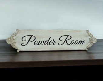 May include: A rectangular, off-white wooden sign with the words "Powder Room" in elegant black script. The sign has ornate, decorative edges and is placed on a dark brown surface.