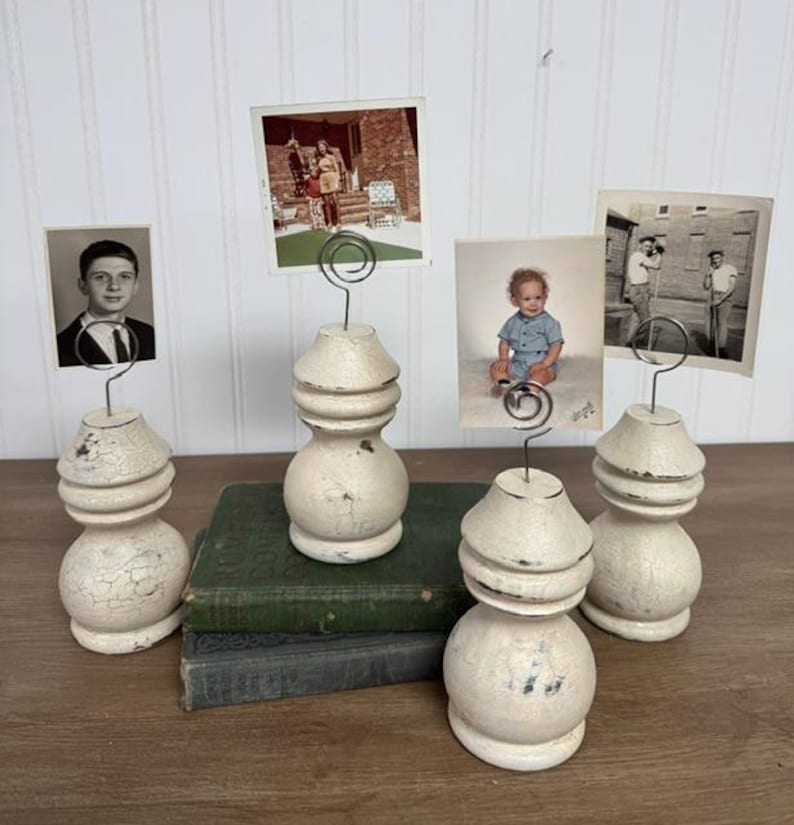 K&ouml;nnte beinhalten: Vier Vintage-Fotohalter mit wei&szlig;em Used-Look, die jeweils ein anderes Foto halten. Die Halter sind aus gedrechseltem Holz gefertigt und stehen auf einem Stapel alter B&uuml;cher. Die Fotos sind in Schwarzwei&szlig; und Farbe.