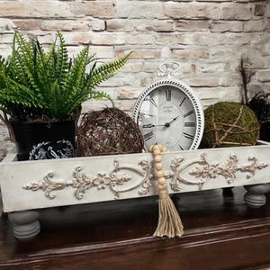 May include: White decorative tray with ornate detailing, featuring a clock, greenery, and a wooden bead garland. The tray has four legs and is sitting on a dark wood surface.