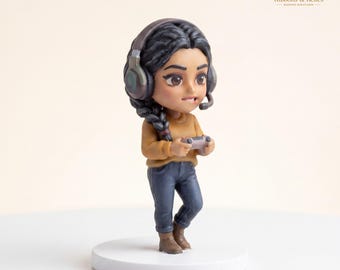 May include: A miniature figurine of a character with dark braided hair wearing a mustard yellow long-sleeve shirt, dark blue jeans, and brown boots. The character wears large over-ear headphones with a microphone and holds a small game controller.
