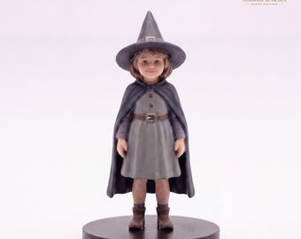 May include: A miniature figurine of a child dressed as a witch, wearing a pointed dark gray hat, a matching dark gray cape, and a light gray dress with a brown belt. The figure stands on a circular black base, featuring a neutral expression.