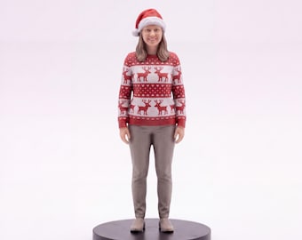 May include: A miniature figurine of a person with shoulder-length brown hair wearing a red and white reindeer-patterned sweater, light brown trousers, and a red Santa hat, standing upright on a circular black base against a plain white background.