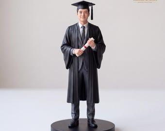 May include: A figurine of a graduate wearing a black academic gown, a matching suit jacket, trousers, and a mortarboard cap with a tassel. The figure holds a white diploma tied with a red ribbon, standing on a circular black base.