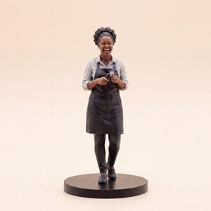 May include: A miniature figurine of a person with dark curly hair, wearing a light-colored button-down shirt, a black apron, and dark pants. The figure is laughing, holding a small tool in their hands, and standing on a circular black base.