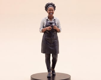 May include: A miniature figurine of a person with dark curly hair, wearing a light-colored button-down shirt, a black apron, and dark pants. The figure is laughing, holding a small tool in their hands, and standing on a circular black base.