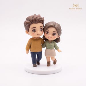 May include: A miniature figurine of two people standing side-by-side on a circular white base. The figure on the left has brown hair, a mustard yellow sweater, and dark blue pants. The figure on the right has brown hair, a sage green shirt, a beige skirt, and brown boots.