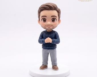 May include: A stylized figurine of a person with brown hair and a light beard, wearing a dark blue cable-knit sweater and light gray trousers. The figure stands on a circular white base with hands clasped together in front of the chest.