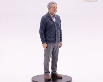 May include: A miniature figurine of an older person with gray hair, wearing a navy blue cable-knit shawl-collar cardigan over a light shirt, gray trousers, and brown shoes. The figure stands on a circular black base against a white background.
