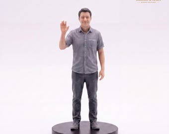 May include: A miniature figurine of a person with short dark hair, wearing a grey short-sleeved button-down shirt and dark grey trousers. The figure stands on a circular black base with one hand raised in a wave gesture, facing forward.