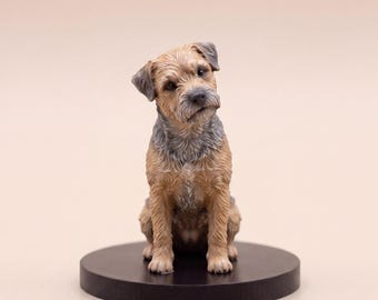 May include: A small, detailed figurine of a Border Terrier dog sitting upright. The dog has a tan and grey wiry coat, dark eyes, and a slightly tilted head. It is mounted on a circular, solid black base, capturing the breed's distinct features.