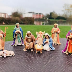 May include: A nativity set featuring hand-painted figurines including a kneeling Joseph, a seated Mary, a baby Jesus in a manger, an angel, three wise men, a shepherd holding a lamb, and two sheep resting on the ground in various colors.