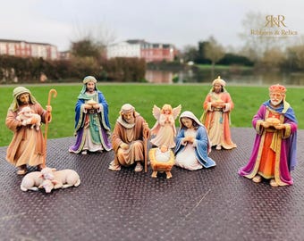 May include: A nativity set featuring hand-painted figurines including a kneeling Joseph, a seated Mary, a baby Jesus in a manger, an angel, three wise men, a shepherd holding a lamb, and two sheep resting on the ground in various colors.