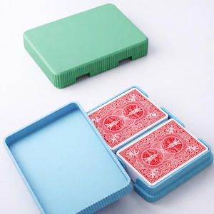 May include: A light blue plastic card case with two decks of playing cards. The cards have red backs with a white design. The case has a green lid and a ribbed design around the edges.