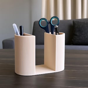 May include: A beige desk organizer with a U-shaped design. The organizer has two compartments, one holding pens and a pen-like object, and the other holding scissors and a pen. The organizer is made of a ribbed material and sits on a dark wooden surface.