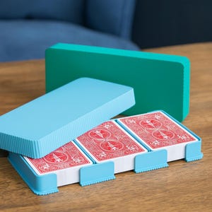 May include: A light blue playing card holder with three compartments, each holding a deck of red playing cards. The holder has a matching light blue lid and a teal green rectangular piece in the background. The holder is on a wooden surface.