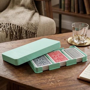 May include: A light mint green plastic playing card holder with a matching lid, containing three decks of cards with blue, red, and black patterned backs. The holder features a ribbed texture on the exterior and is designed to keep the card decks organised.