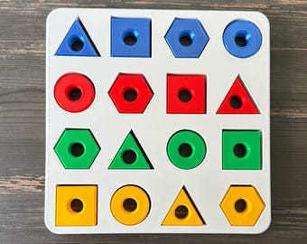 Op de afbeelding: Een witte houten vormsorteerder met diverse gekleurde geometrische vormen. De vormen omvatten blauwe driehoeken, vierkanten en zeshoeken; rode cirkels, zeshoeken en vierkanten; groene cirkels, driehoeken en vierkanten; en gele vierkanten, cirkels en zeshoeken.