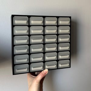 May include: A black storage organizer with 30 small, clear drawers. Each drawer has a white label area. The organizer is held by a hand against a white wall. Ideal for small parts storage.